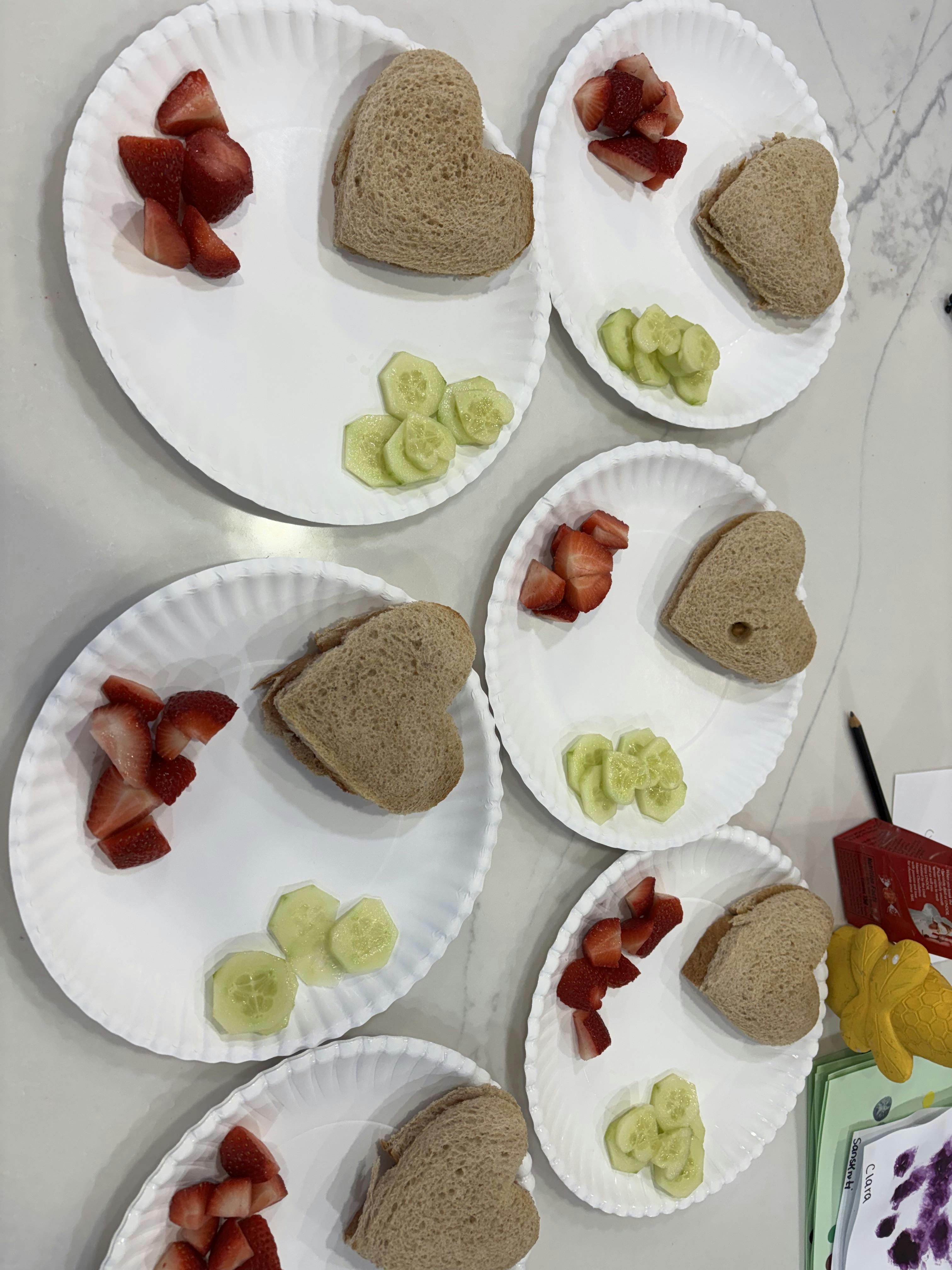 Multiple white paper plates with heart-shaped sandwiches, sliced strawberries, and cucumber