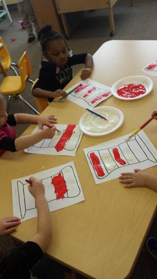Children painting red and white stripes on paper at a table