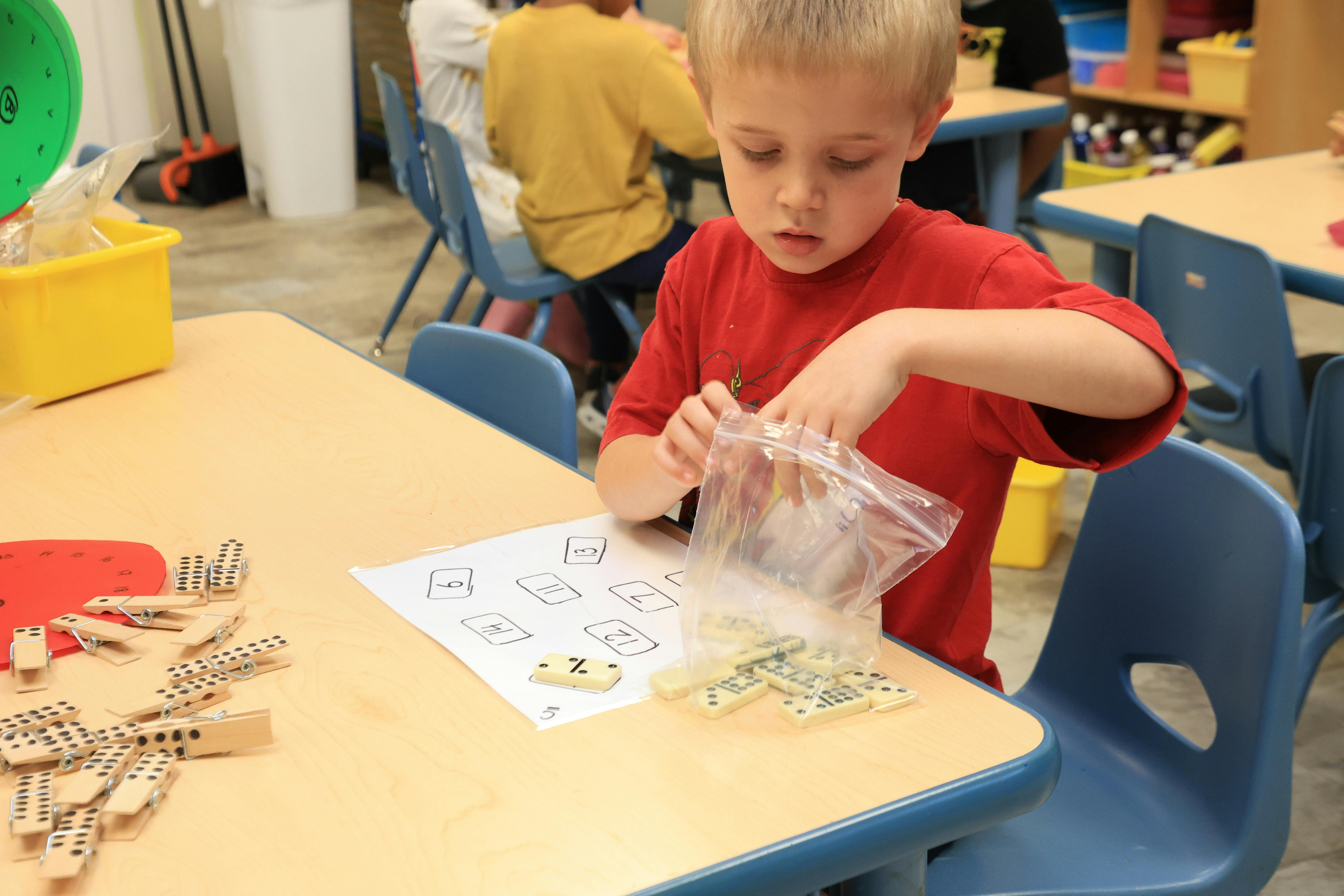Child at a table with a worksheet, taking items from a plastic bag