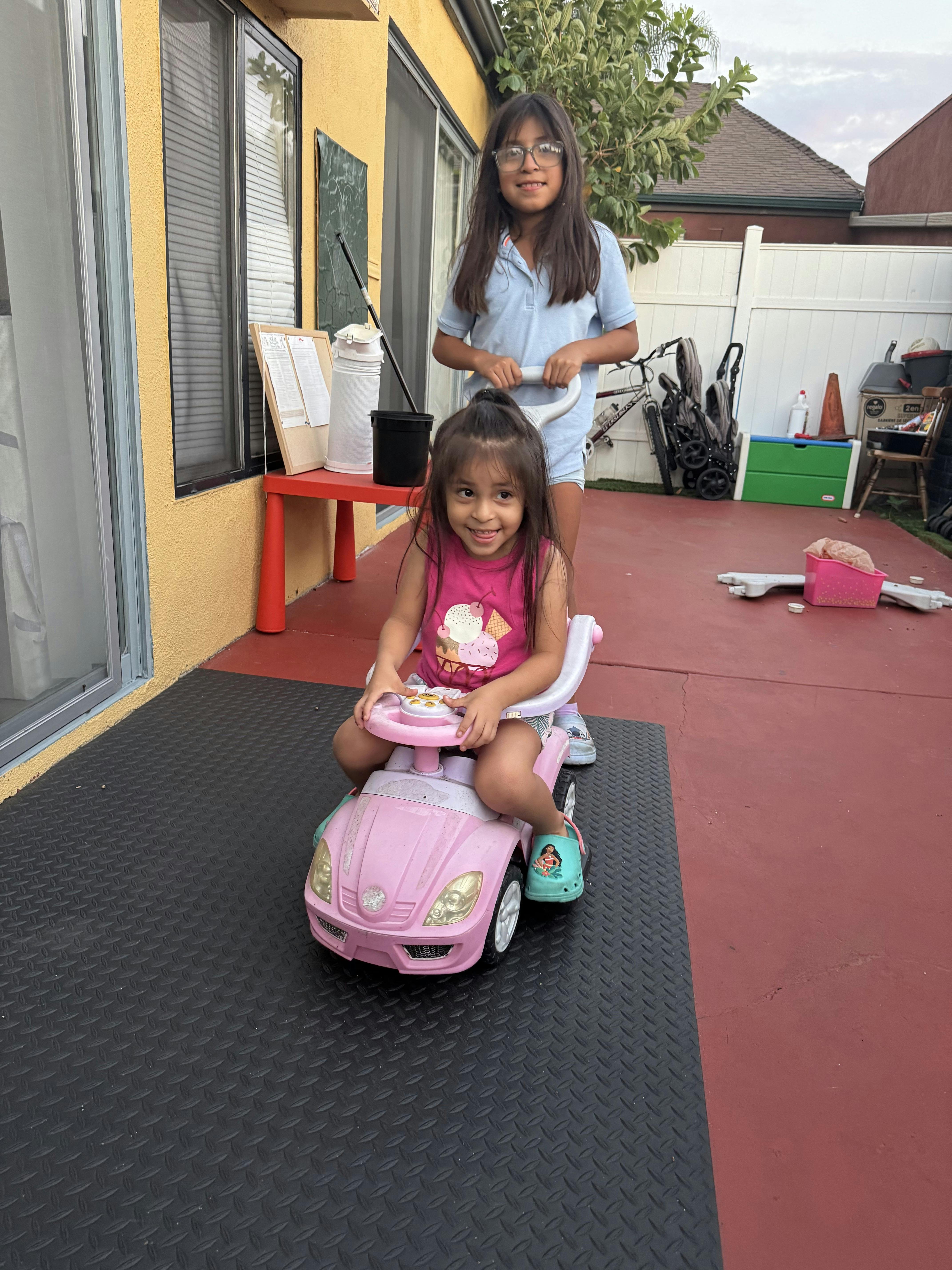 A teen pushing a child on a toy car in a backyard