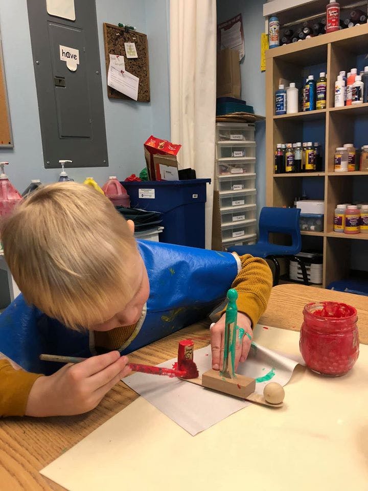 Child in a blue smock painting a wooden object at a table