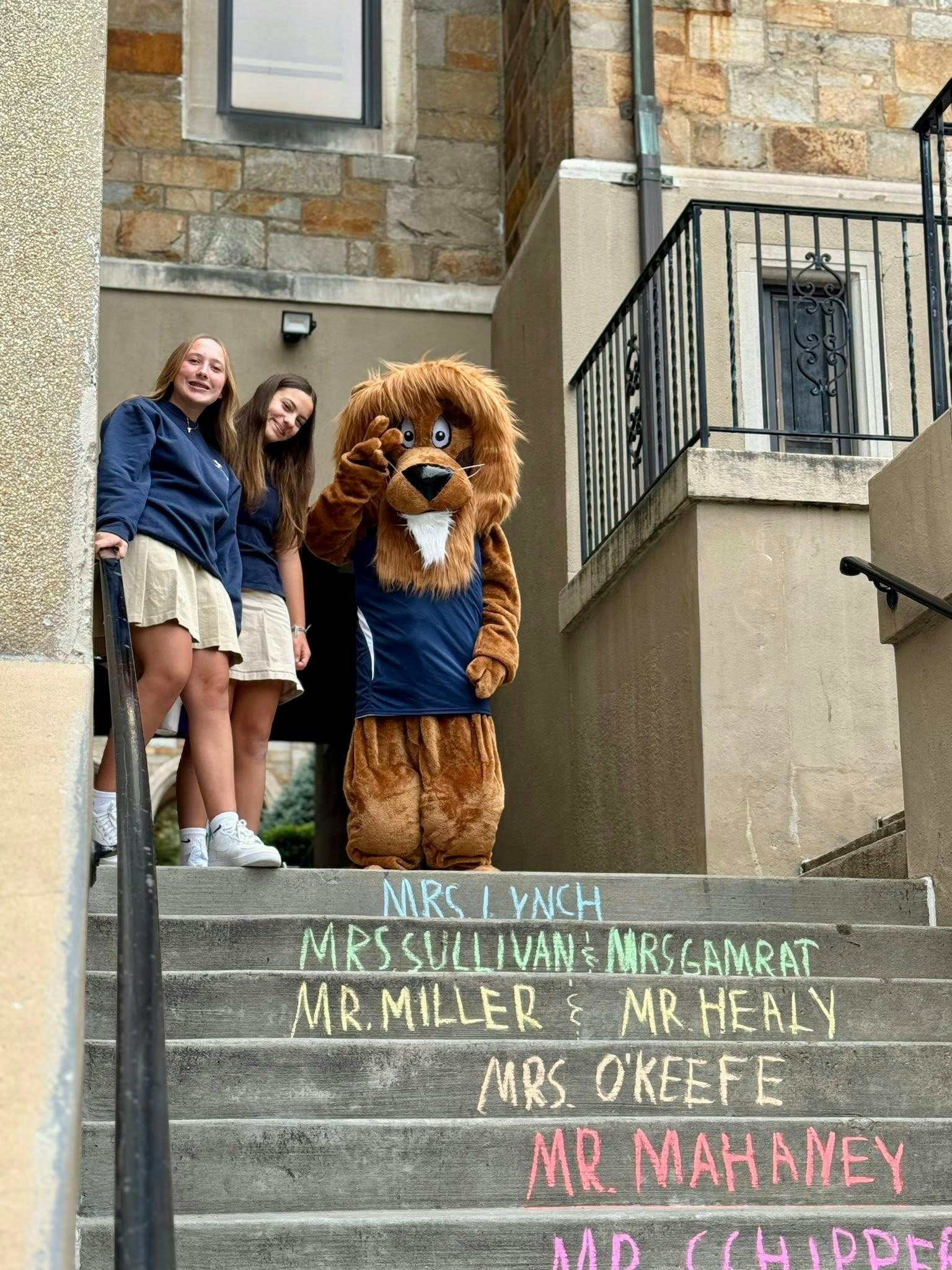 Two teens and a lion mascot on stairs with names written in chalk