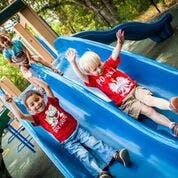 Two children sliding down a blue slide, another child at the top of the slide