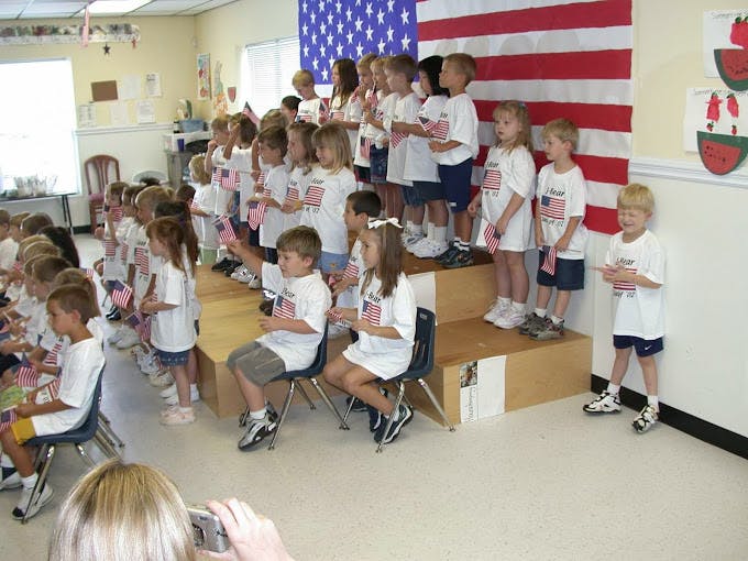 Children in white shirts standing and sitting in a room with an American flag