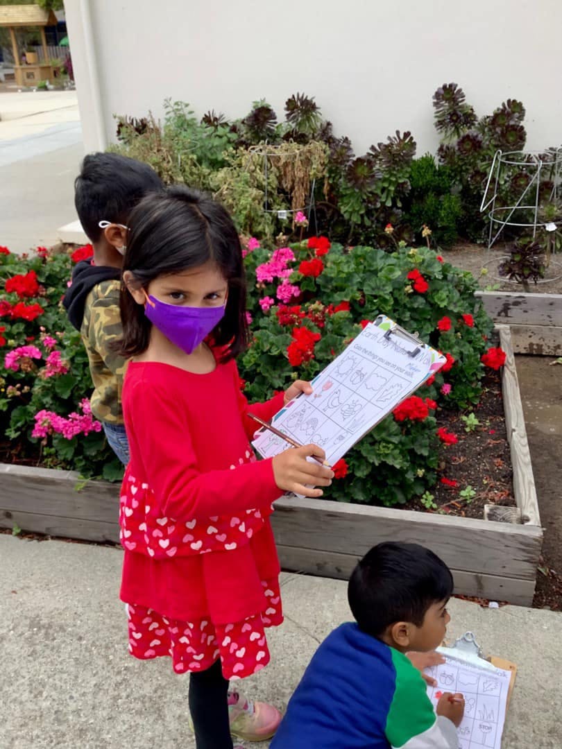 Child in a purple mask holding a clipboard with other children in a garden