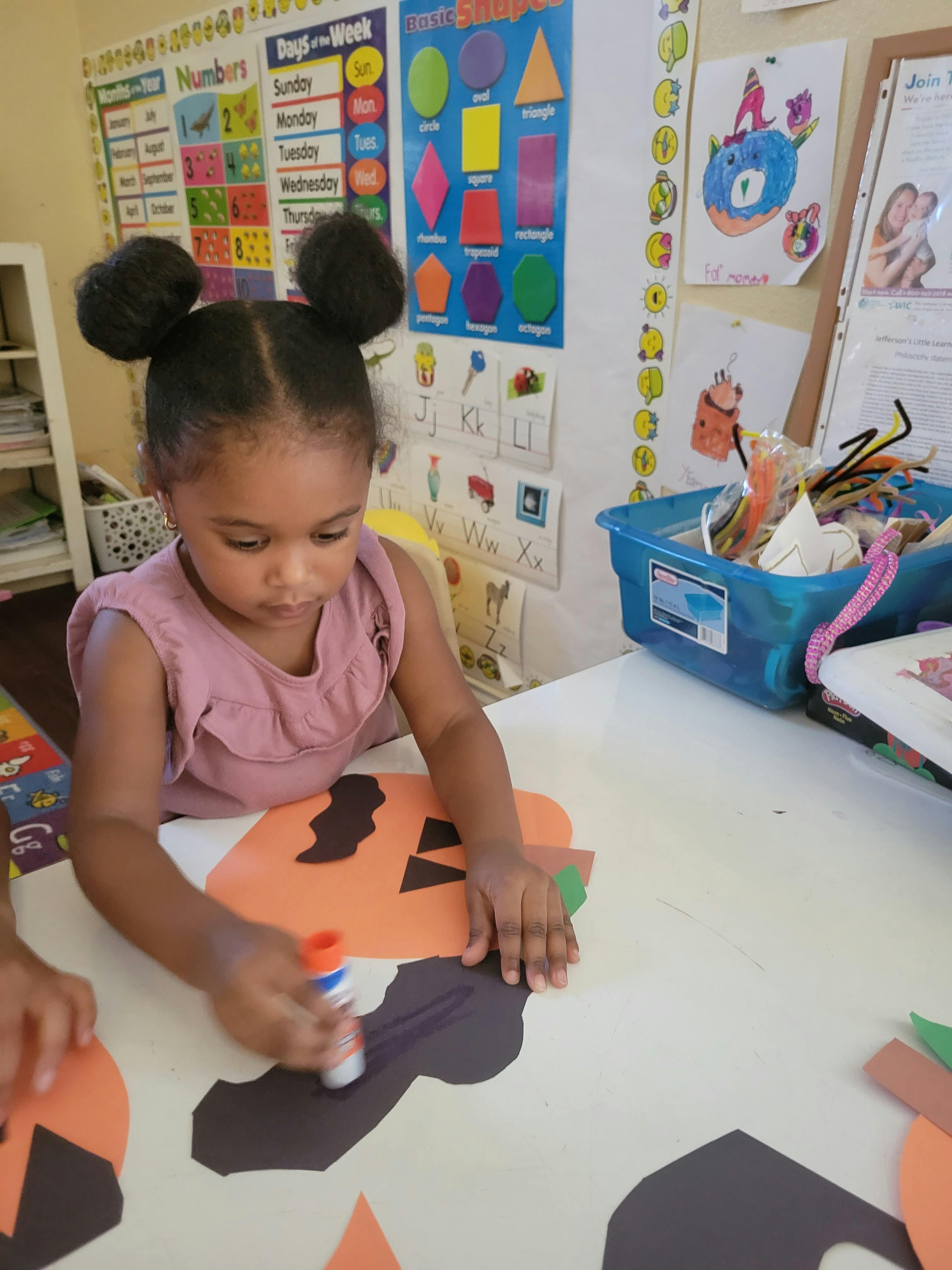 Child gluing paper cutouts onto an orange paper shape