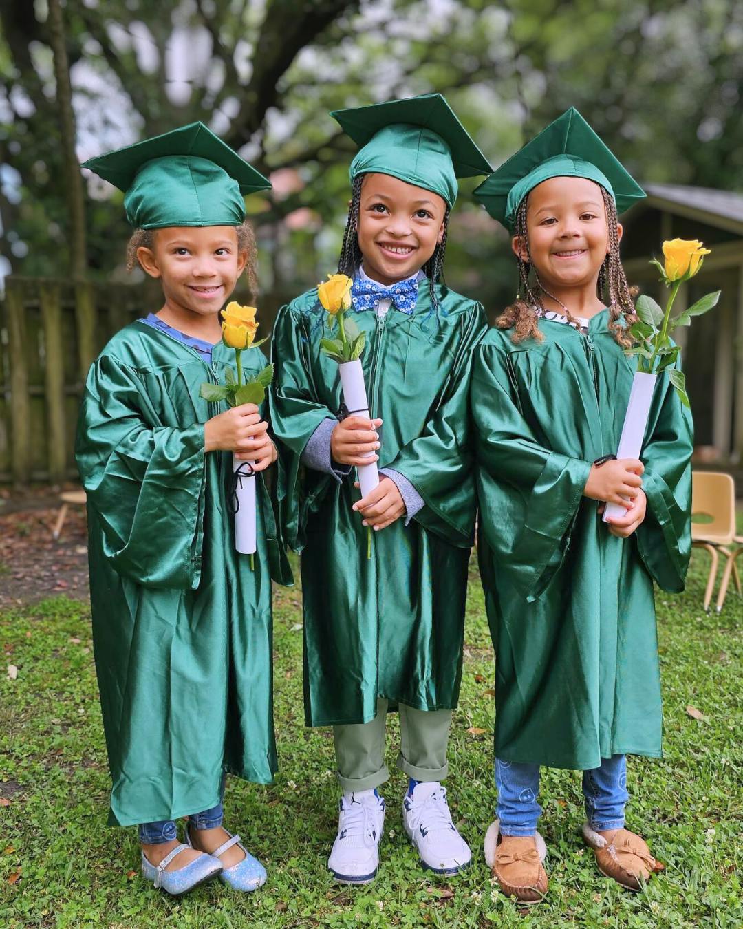 Three children in green graduation caps and gowns holding yellow roses and diplomas
