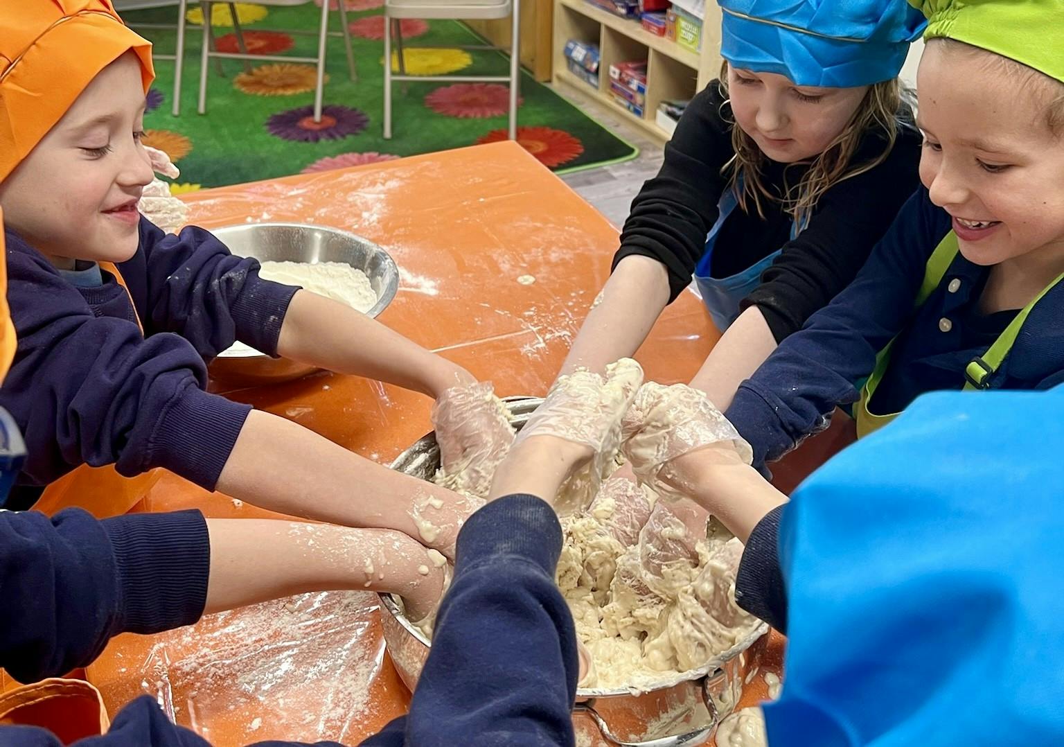 Several children in aprons and chef hats mixing dough in a large bowl