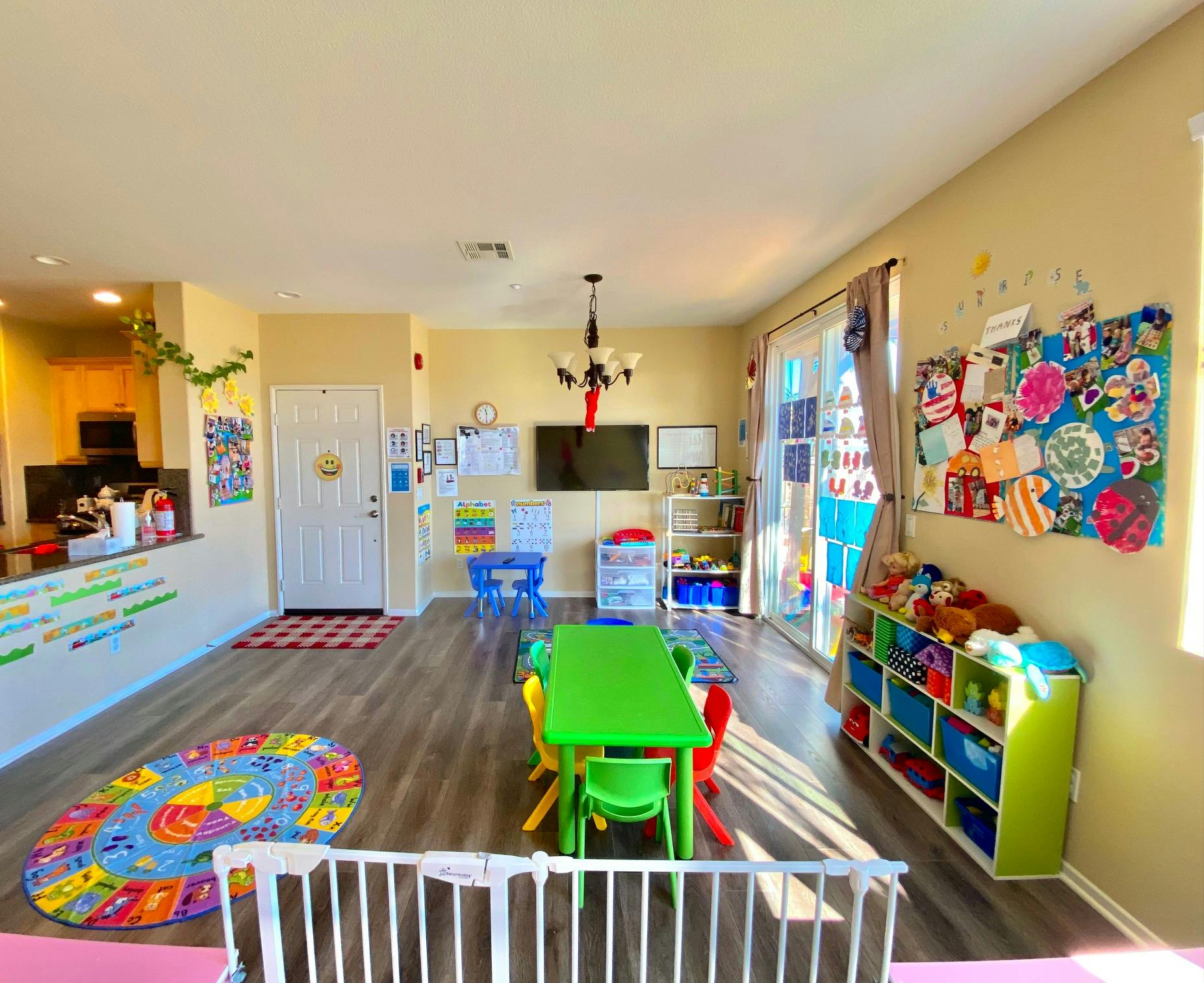 A brightly lit room with a green table and chairs, colorful rug, and children's artwork