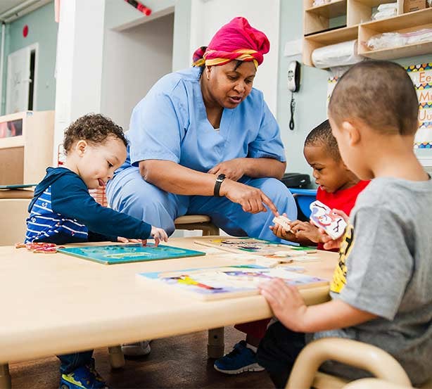 Person in scrubs pointing at a table with three children playing with puzzles
