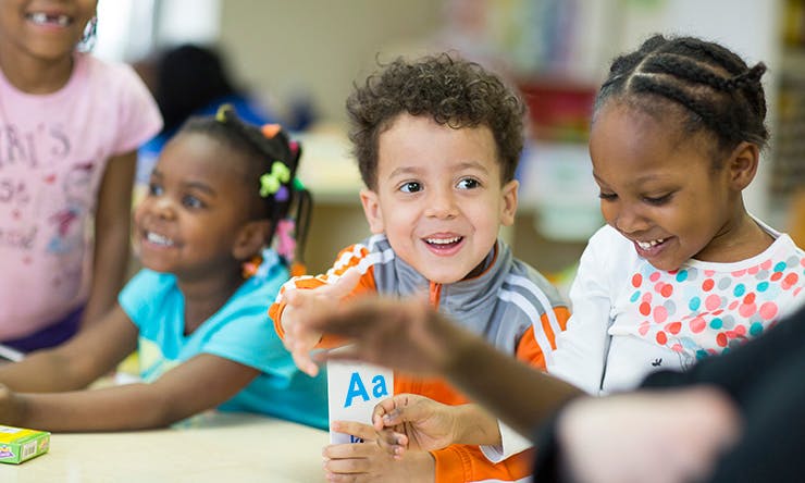 Children in a classroom, one holding a card with "Aa"