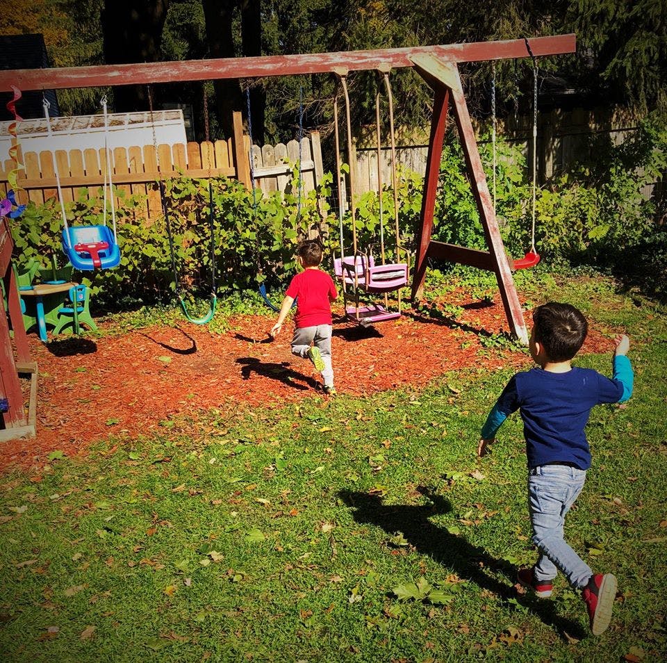 Two children running on grass and wood chips near a swing set
