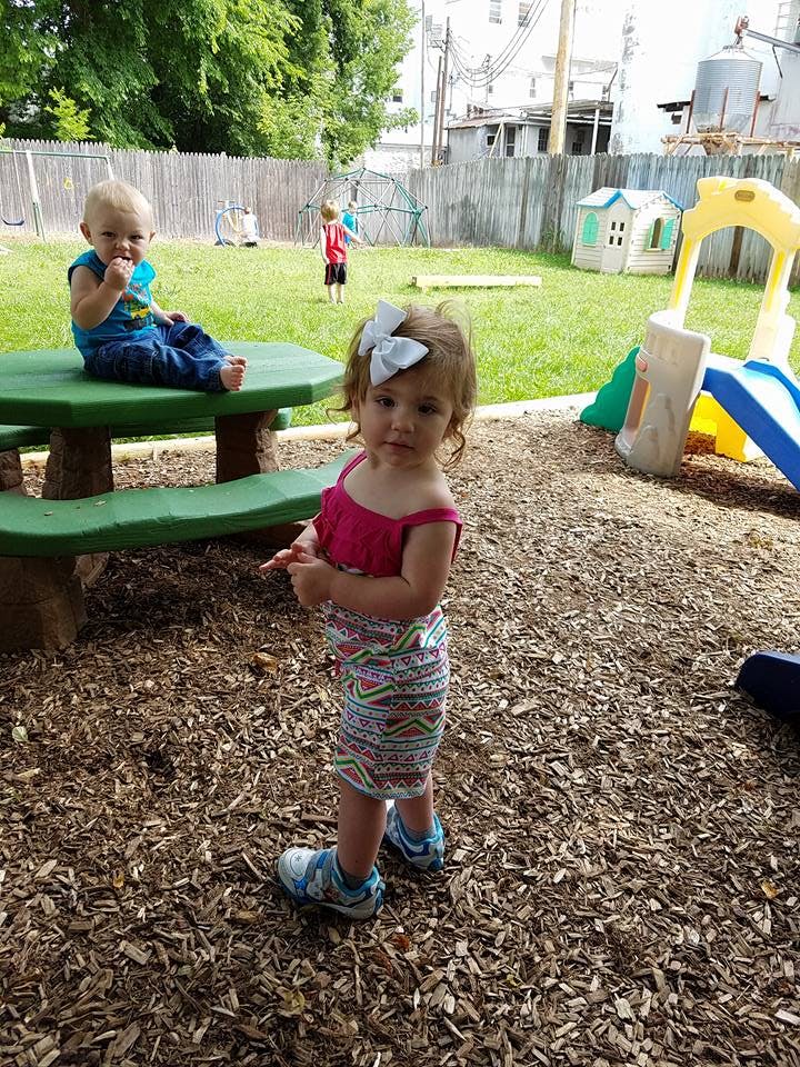 Child standing on wood chips in a playground, another child on a picnic table