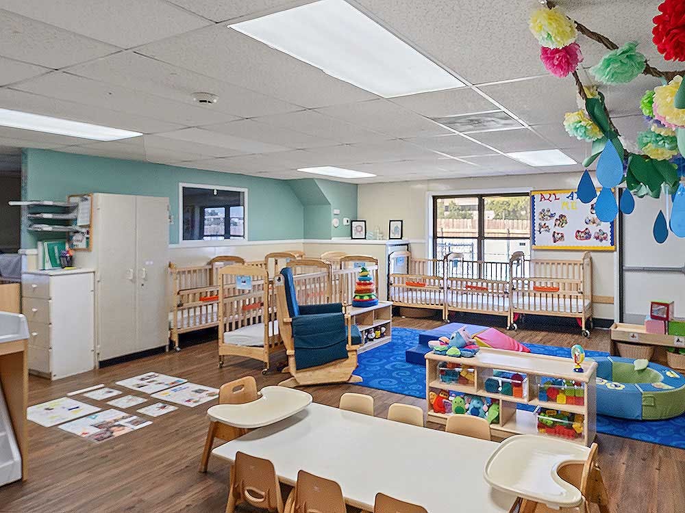 A classroom with cribs, a long table with chairs, and a play area