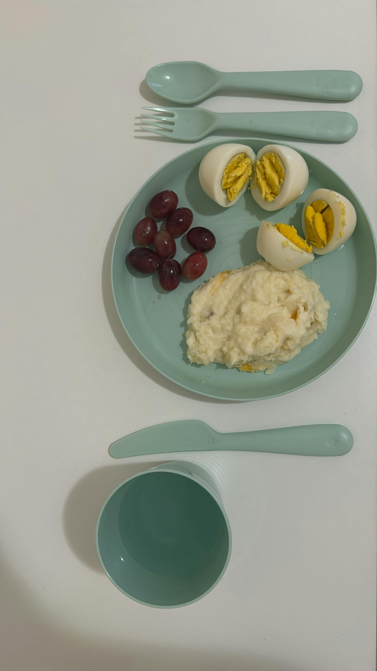 Plate with grapes, hard-boiled eggs, and mashed food, with cutlery and a cup