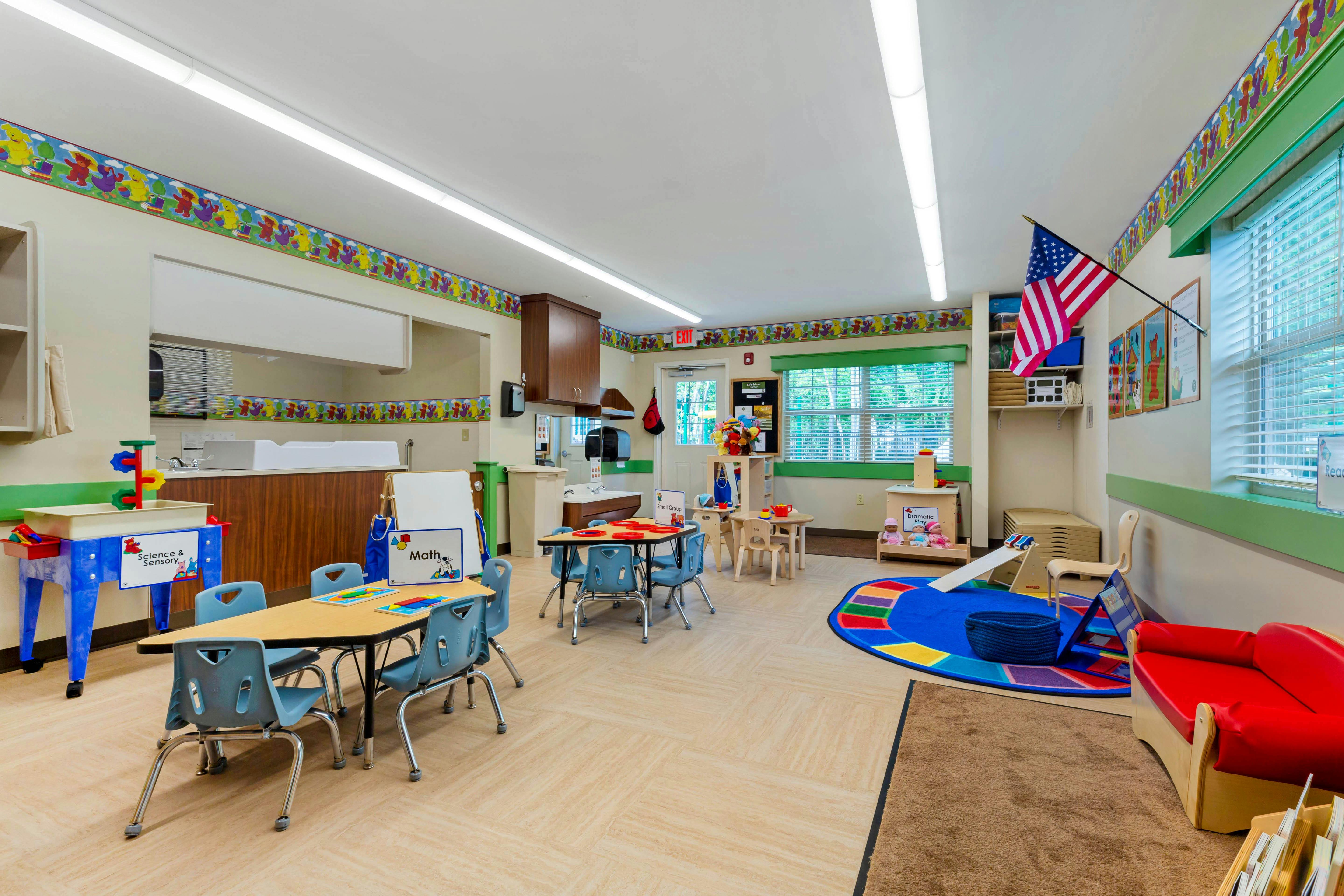 Classroom with child-sized tables and chairs, a play area with a rug, and a sink