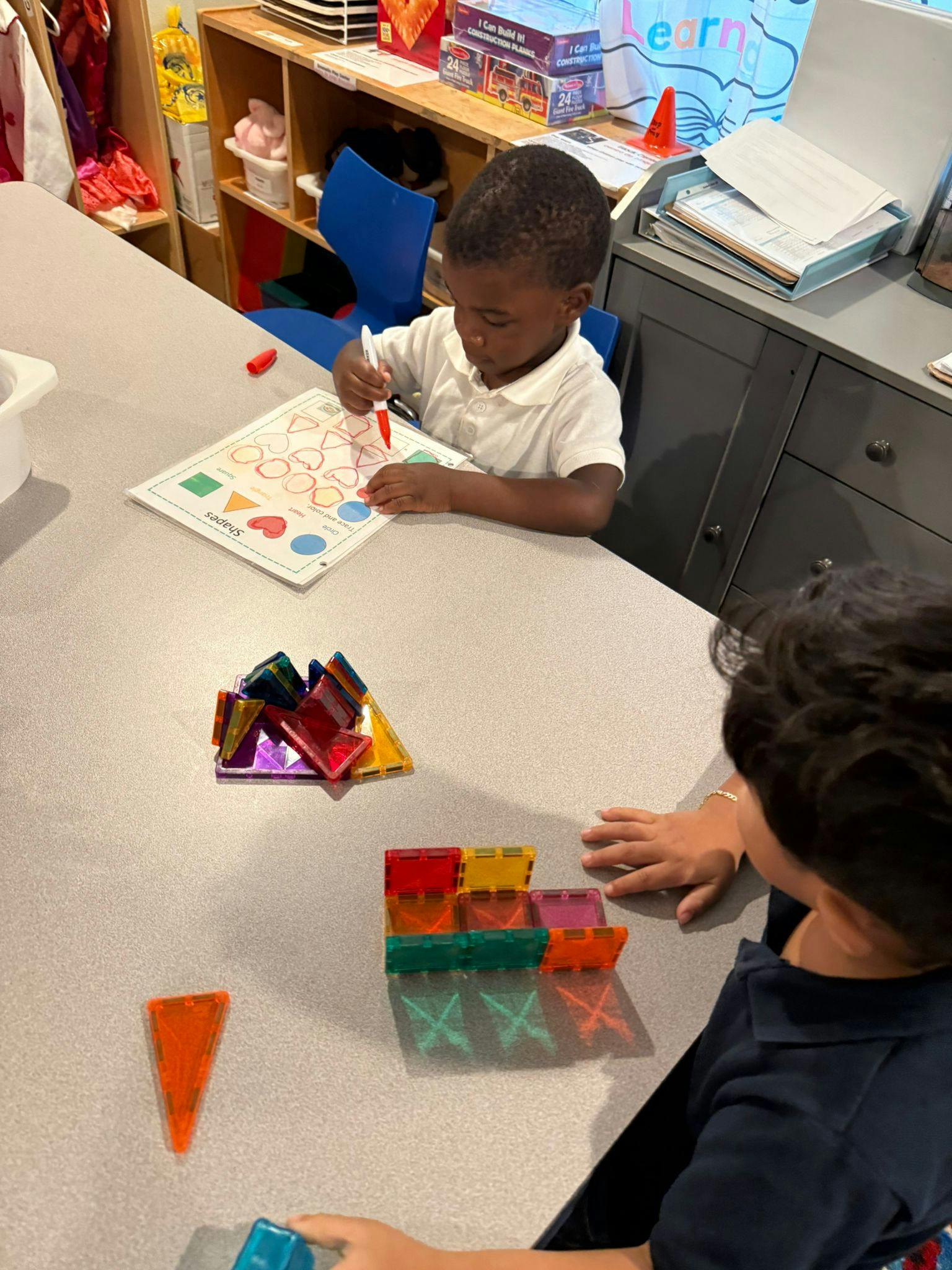 Child drawing on paper at a table, another child playing with magnetic tiles