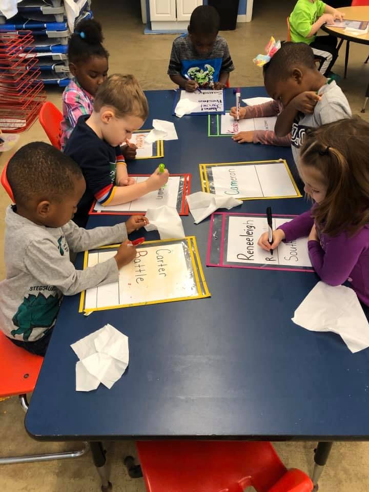 Children writing on papers at a table in a classroom
