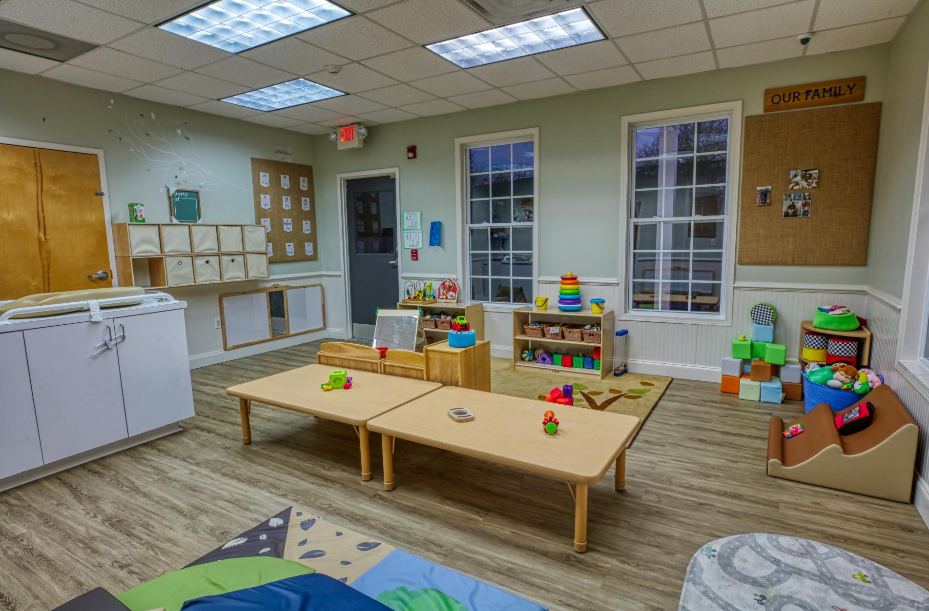 A brightly lit classroom with small tables, toys, and changing station