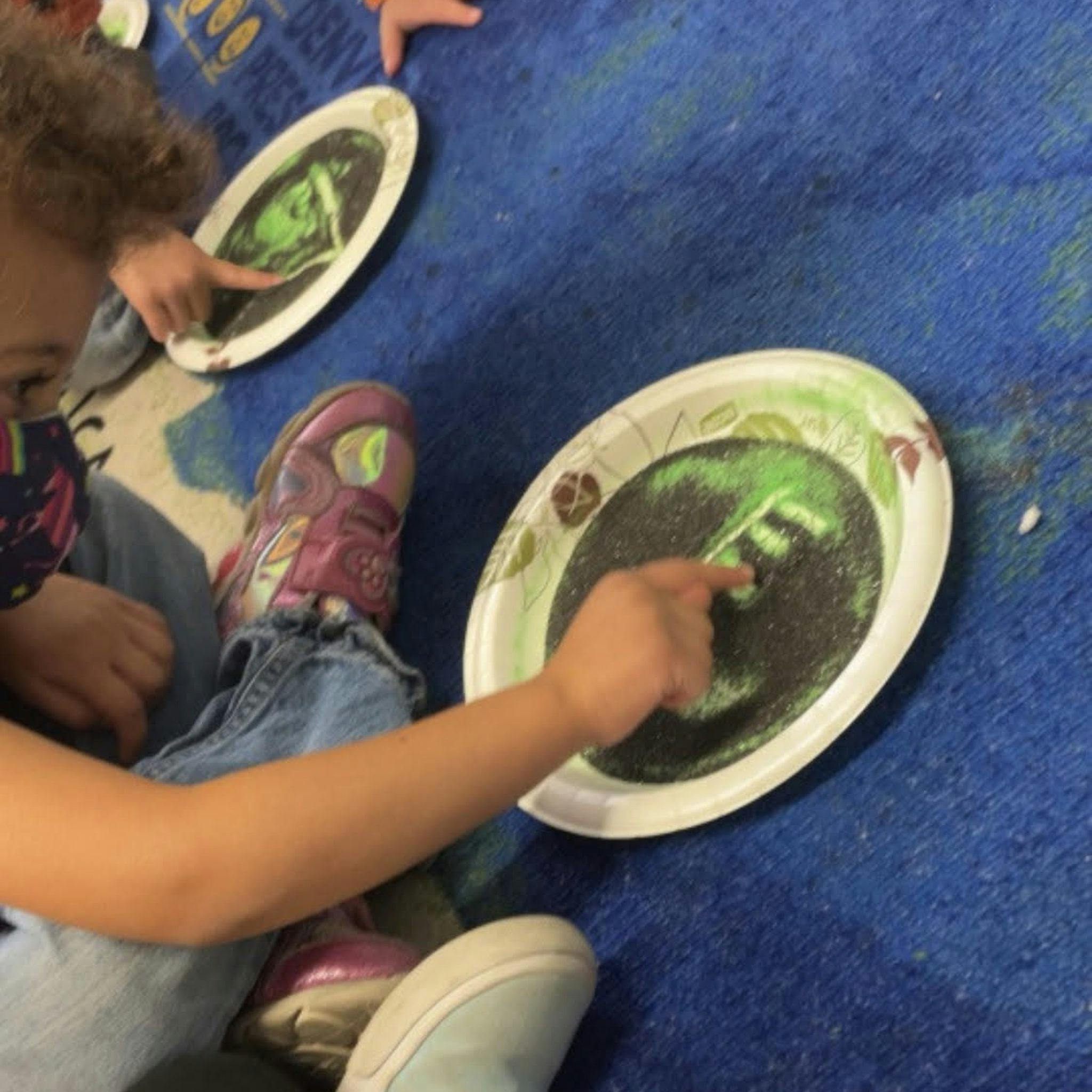 Child's hand pointing at green and black sand on a paper plate