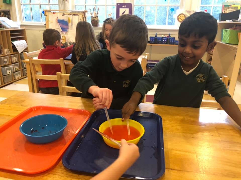 Two children mixing liquid in a bowl at a table in a classroom