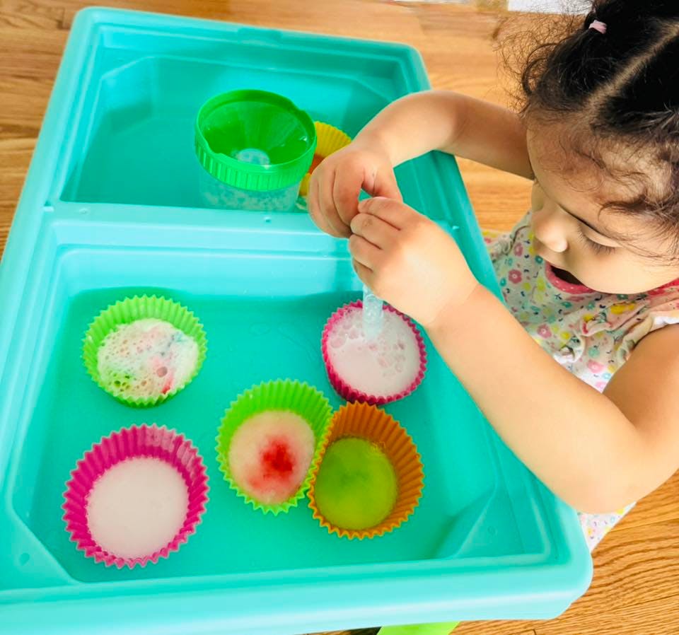 Child playing with colorful cupcake liners filled with foamy liquid in a teal bin