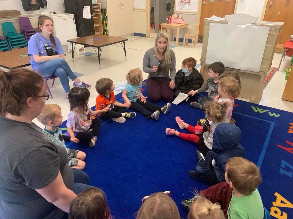 Adults and children sitting in a circle on a blue rug in a classroom