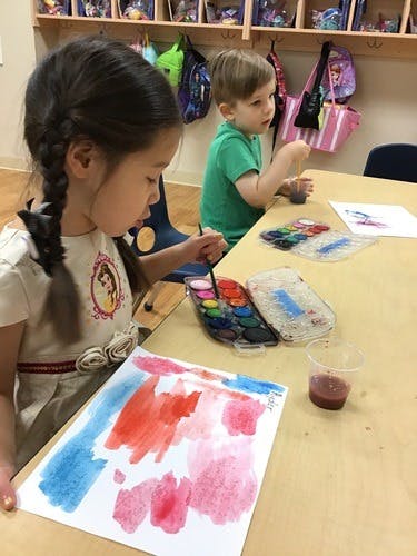 Child painting with watercolors at a table, another child in background