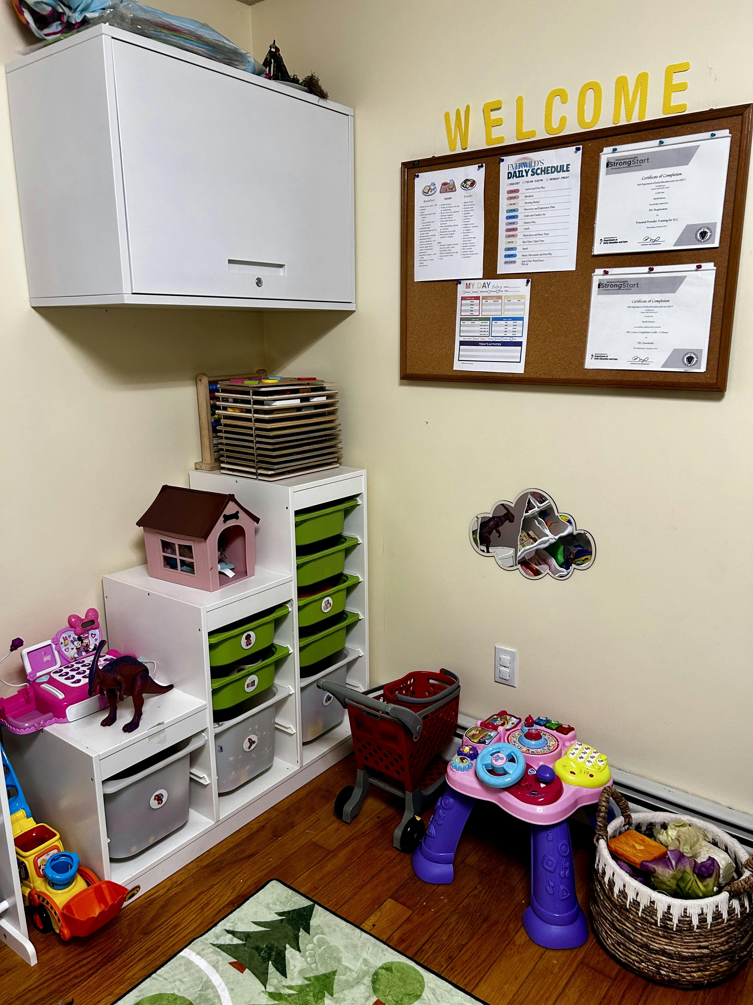 Childcare room with toys, storage, a bulletin board, and a "WELCOME" sign