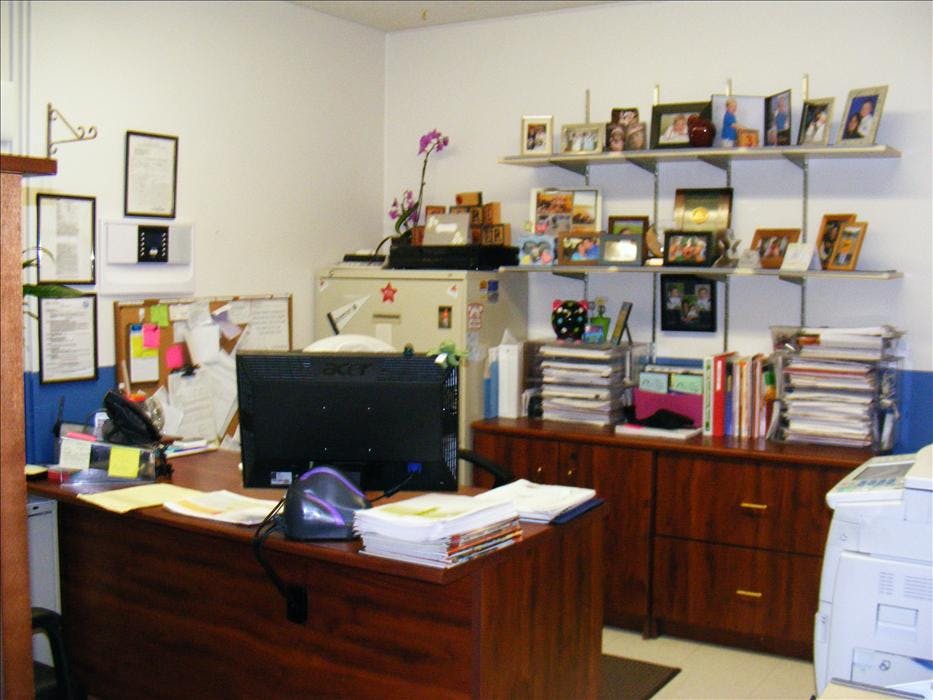 A cluttered office desk with a computer monitor, papers, and shelves full of framed photos