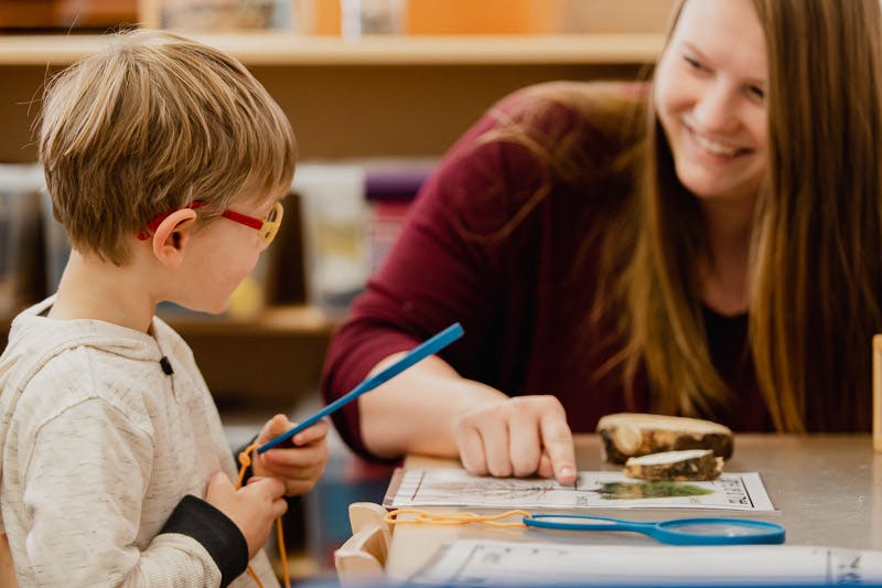 Child in glasses holding a stick and person pointing at a paper