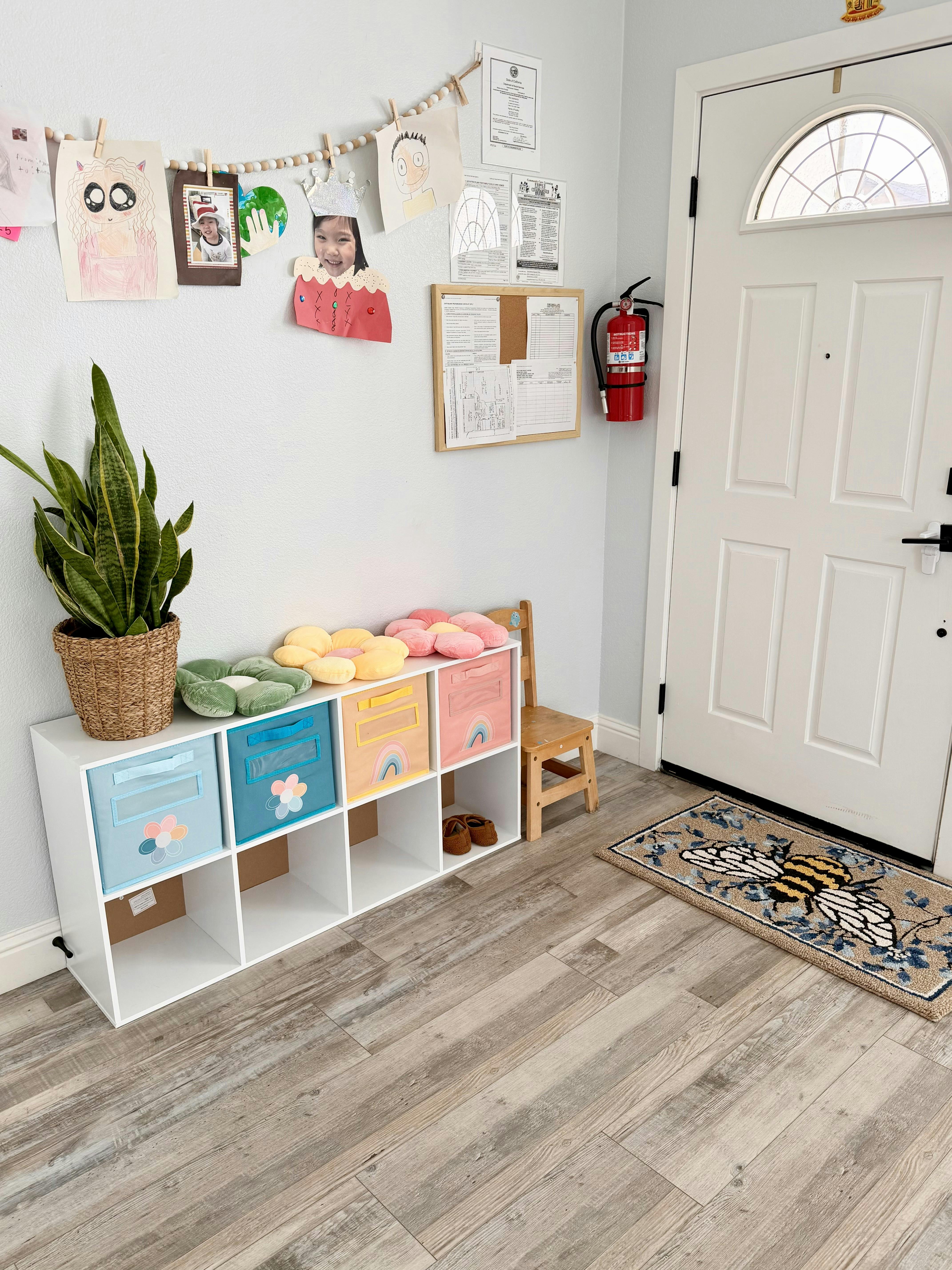A home entryway with kids' artwork, a storage shelf with bins, a plant, and a white door