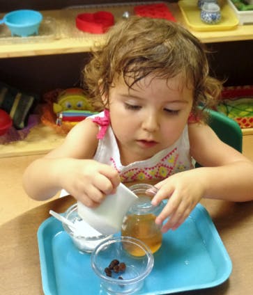 Child pouring liquid from a small pitcher into a jar on a blue tray