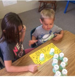 Two children playing a game with cups on a yellow mat at a table
