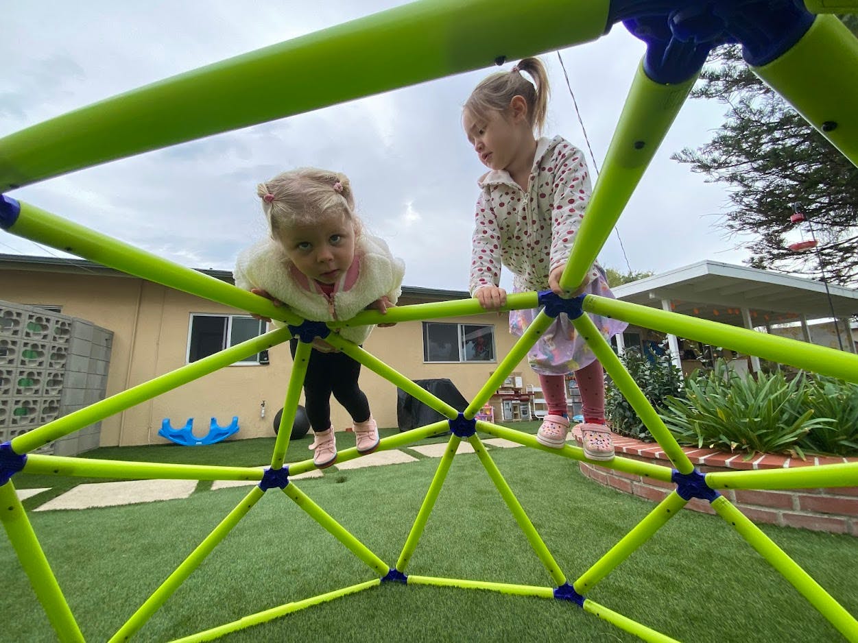 Two children climbing on a green geodesic dome climber on a grassy play area