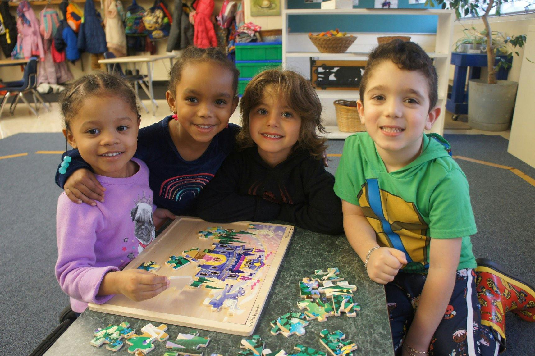 Four children smiling while doing a puzzle in a classroom