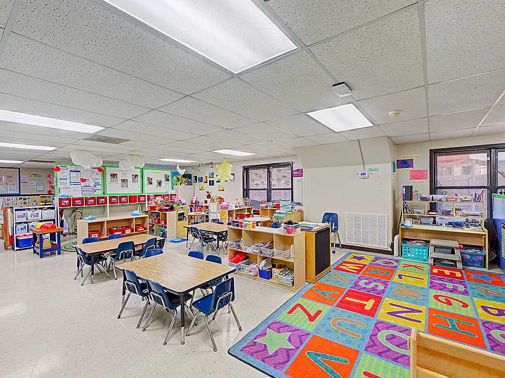 Empty classroom with small tables and chairs, a colorful alphabet rug, and shelves
