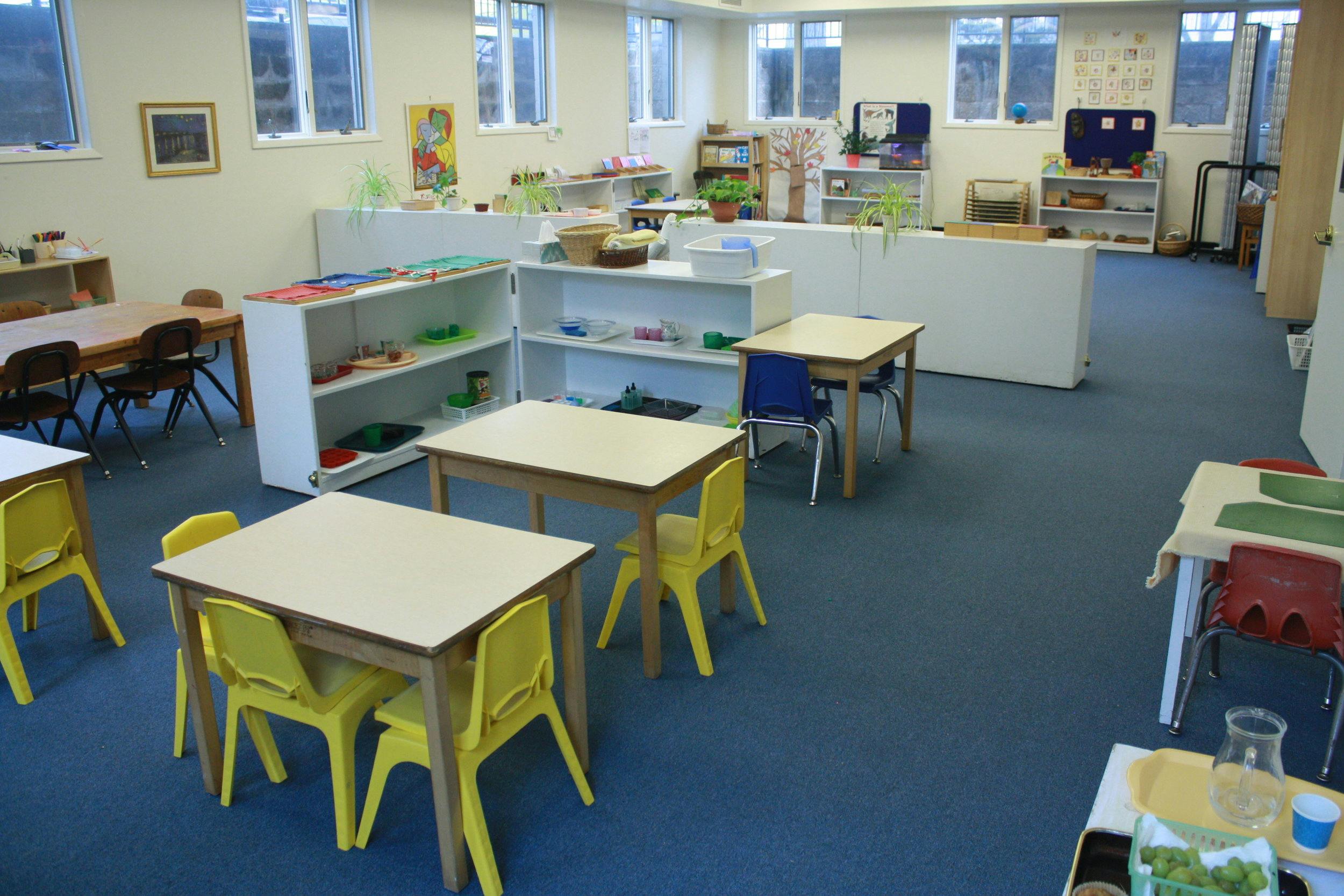 Classroom with small tables and chairs, shelves with learning materials