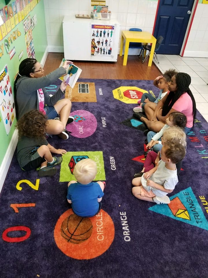 Adults and children sitting on a rug, one adult reading a book to the group