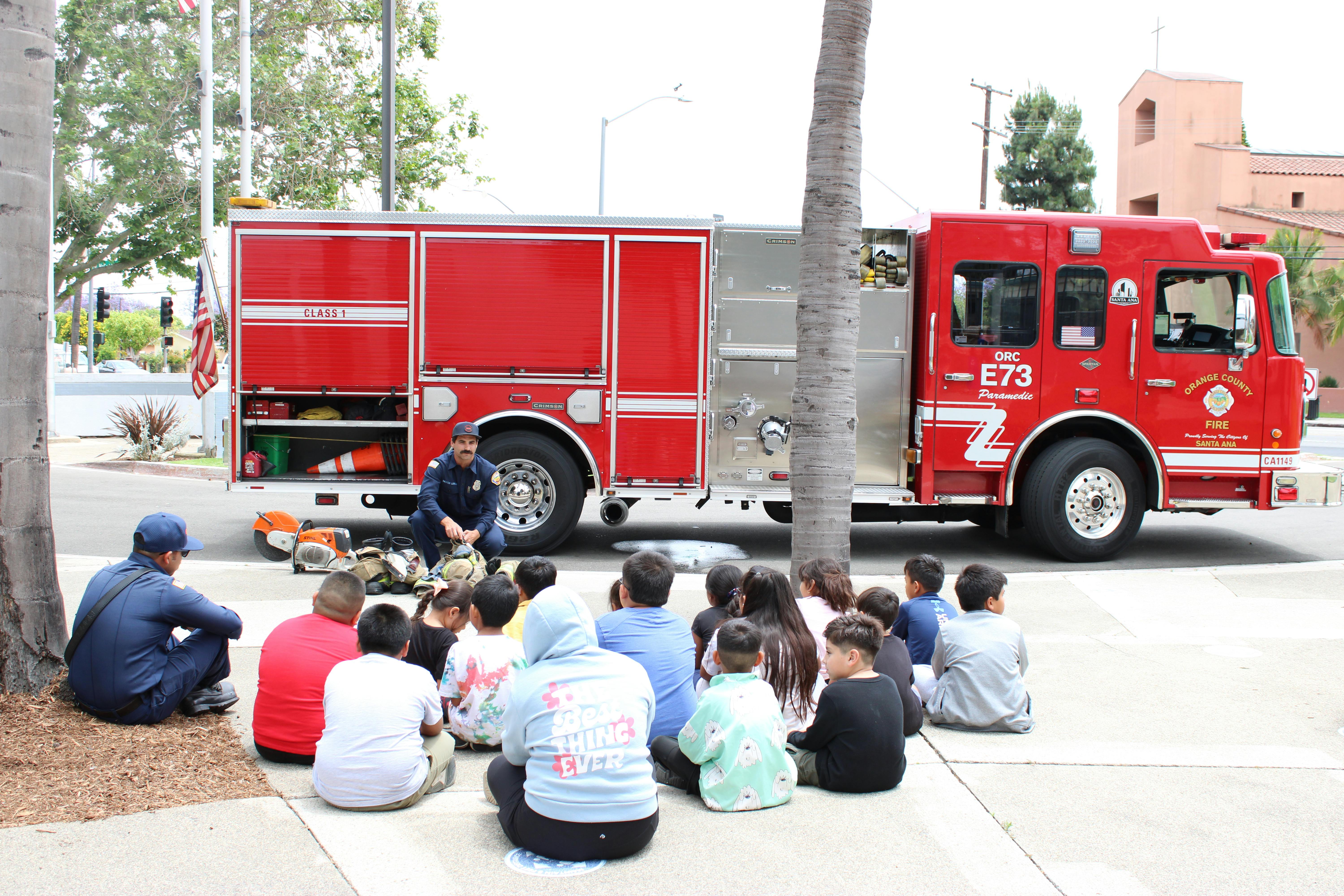 Firefighter crouching in front of a fire truck speaking to children sitting on the ground