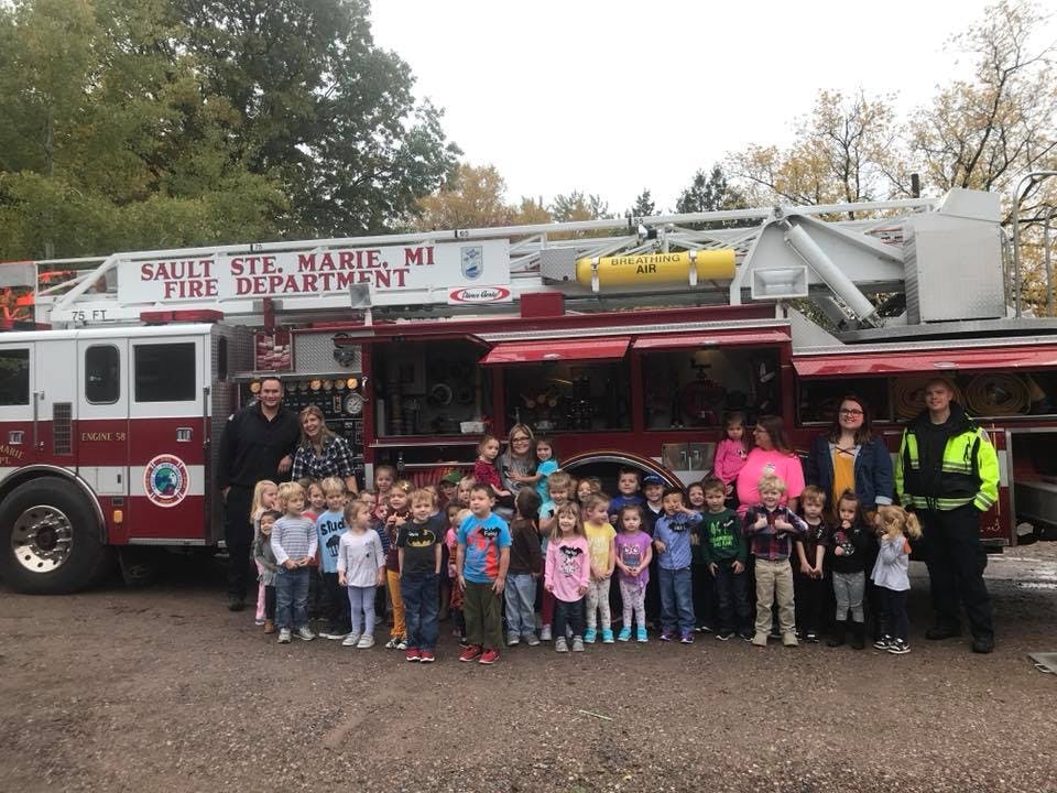 People and children standing in front of a fire truck with "Sault Ste. Marie, MI Fire Department" on it