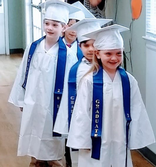 Children in white graduation gowns and caps with blue sashes that say "GRADUATE"