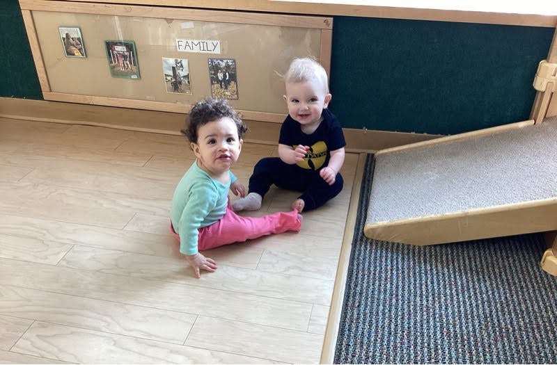 Two infants sitting on a wooden floor in a classroom