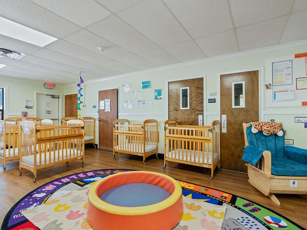 A room with several cribs, a blue rocking chair, and a circular play mat