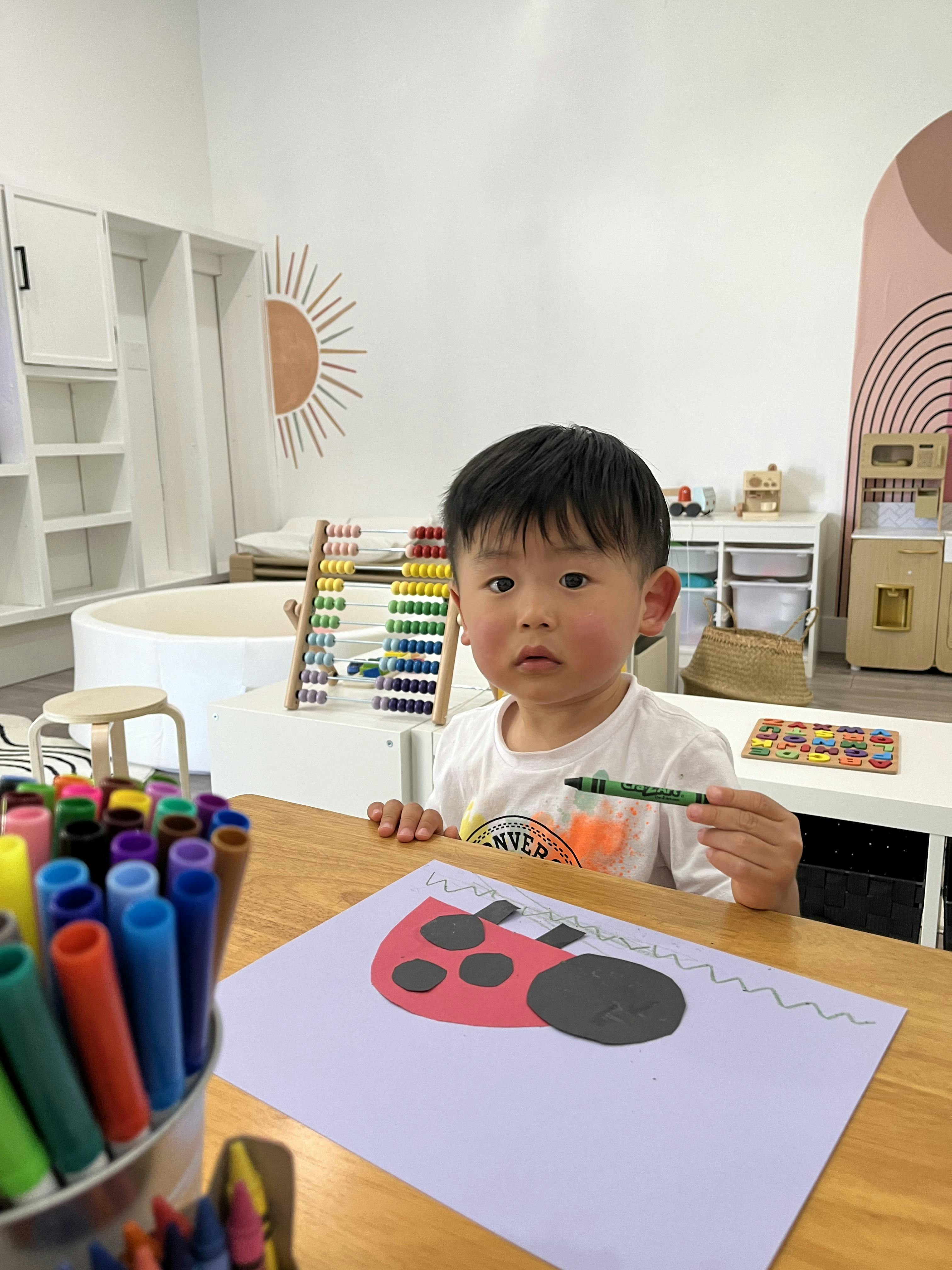 Child at a table holding a green crayon, next to a ladybug craft