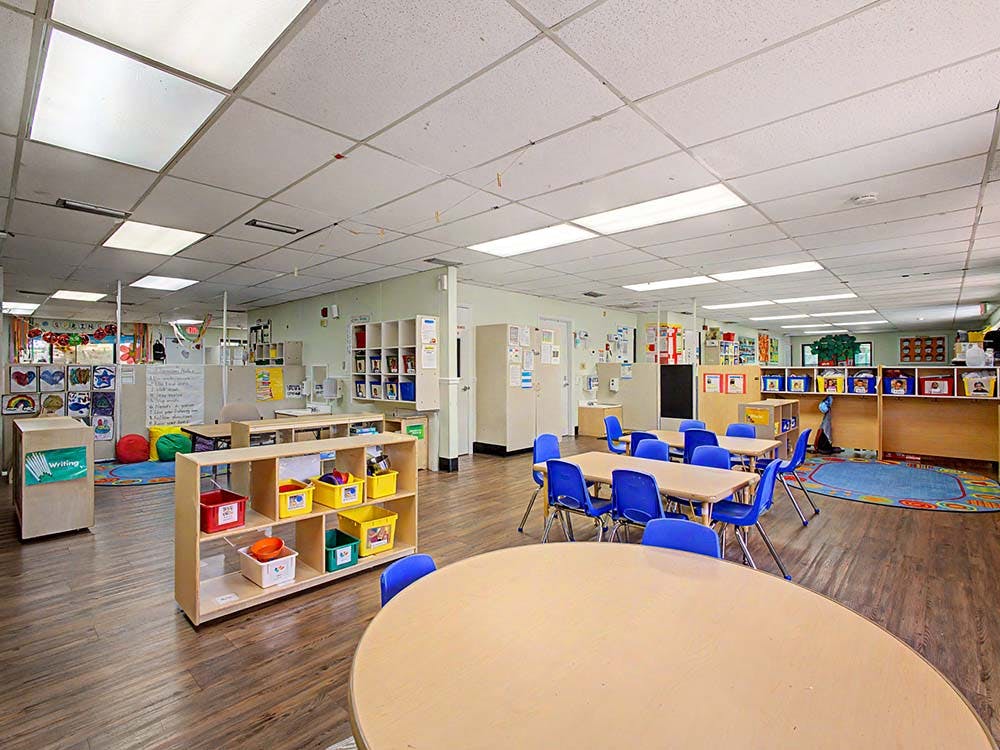 Empty classroom with small tables and chairs, shelves with storage bins, and artwork