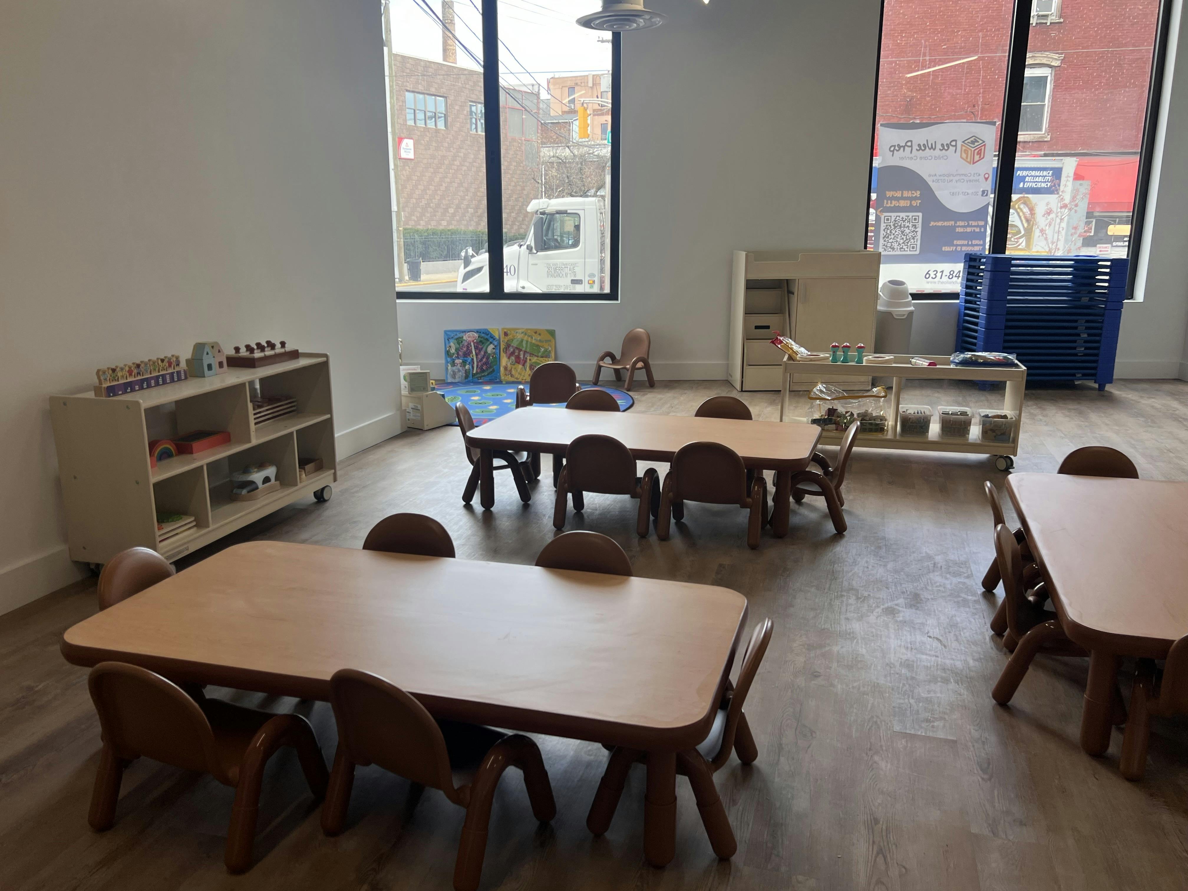 Classroom with several small tables and chairs, shelves with toys, and large windows