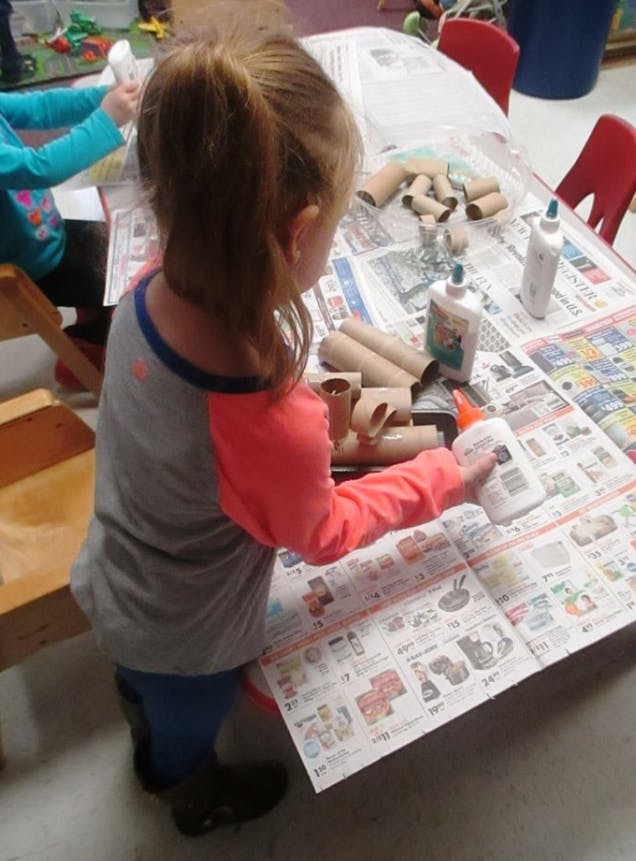 Child holding a glue bottle over newspaper on a table with cardboard tubes