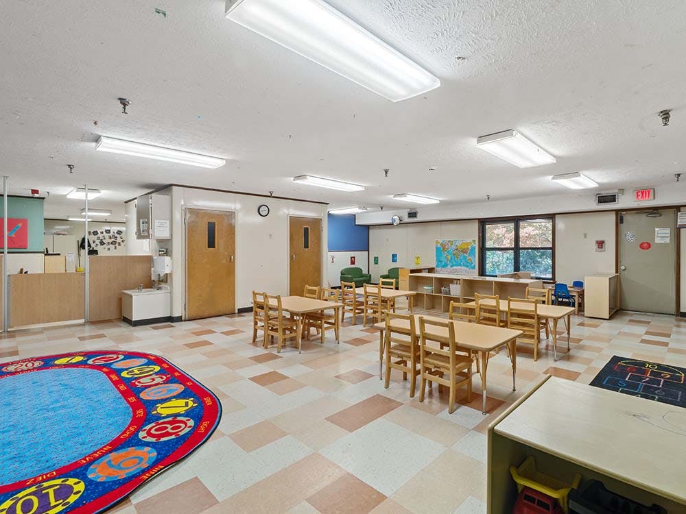 Empty classroom with small wooden tables and chairs, a play rug, and window