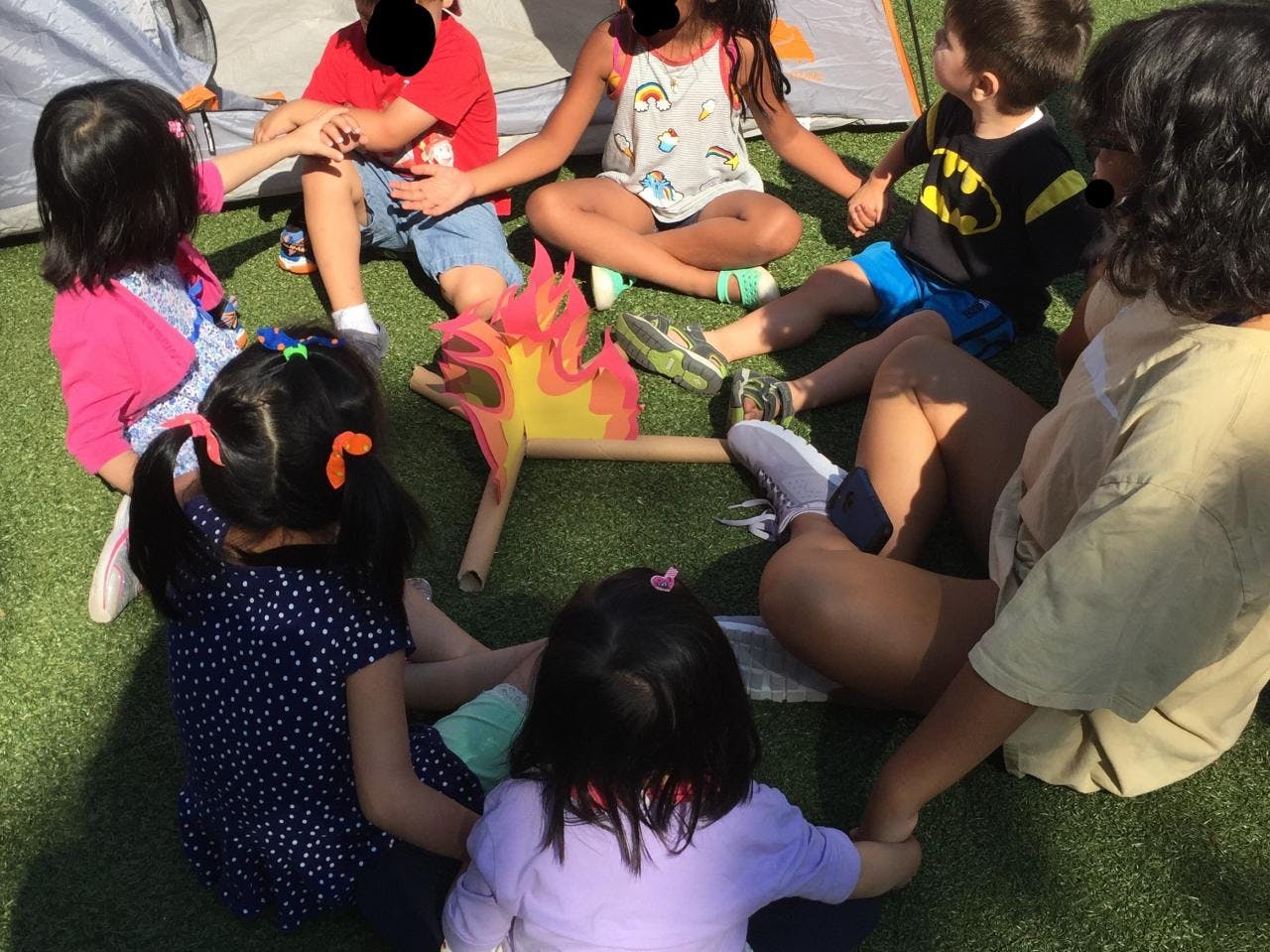 Children and adults sitting in a circle on grass around a pretend campfire